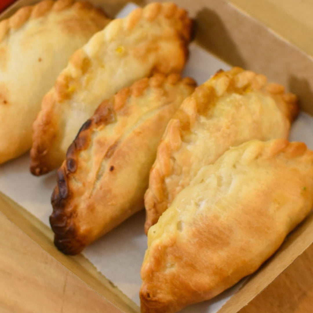 A box of freshly baked Argentinian-style empanadas with various fillings, displayed on a wooden board with a knife and a sprig of parsley nearby.