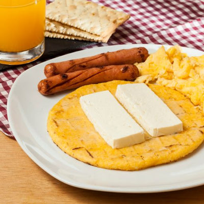 Yellow Corn Colombian Arepa topped with Paisa Cheese, accompanied by two sausages and scrambled eggs, beautifully presented on a white plate