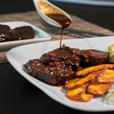 Delectable black caramelized beef with a tantalizing sauce being poured with a spoon, served alongside perfectly ripened plantains