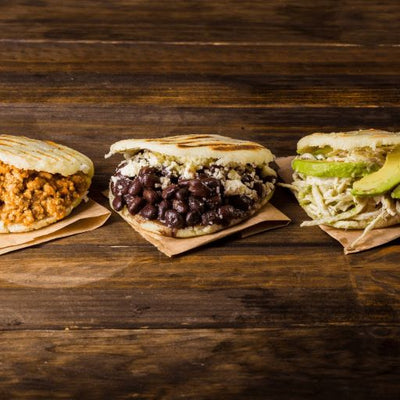 Three Arepas on a wooden board with different fillings. From left to right: 1. minced beef. 2. Black beans and white cheese. 3. Shredded chicken, white cheese and avocado.